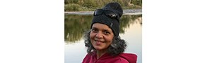 A smiling woman in a gray winter hat and a red shirt, with sunglasses perched on her head, stands in front of a green river with trees in the background.