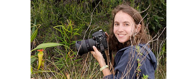 Smiling woman with long brown hair holds camera pointed toward yellow and red flower while crouched on the ground in a grassy field