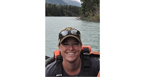 Smiling woman wearing baseball cap with sunglasses perched on top, with water, trees, and high mountains behind her