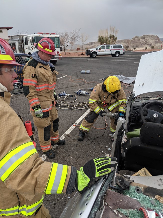 Auto Extraction Three structural firefighters stand near a crashed vehicle, one firefighter is using a tool to get inside the vehicle.