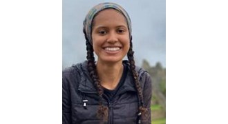 Avani Fachon Fachon wearing a bandana, braids, and a black puffy vest, outside at a scenic vista smiling at the camera.