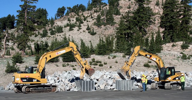 Two frontloaders load rocks into large buckets in front of a rock and tree-covered mountain