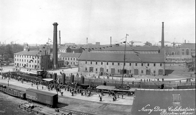 B22Wimg2_historical A black and white photo giving a view of the Charlestown Navy Yard from above. In the midground is a dry dock with a navy ship. Behind it and to the left is Building 22. Several industrial buildings are throughout the Navy Yard.