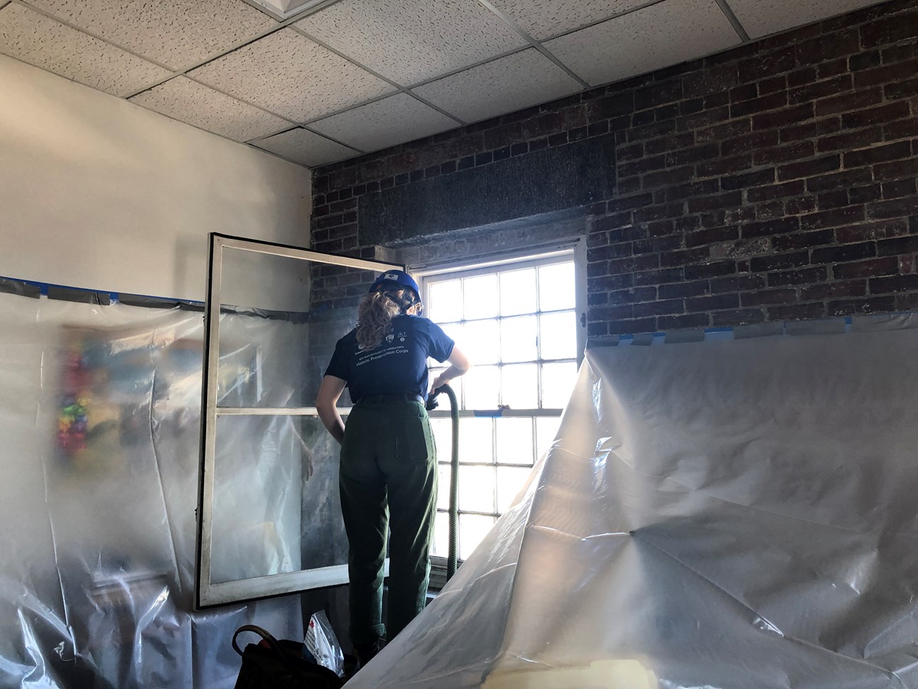 A Historic Preservation Corps member standing on a platform in front of a window, vacuuming it in preparation for removal. Around them the furniture and walls are covered with semi-clear plastic sheeting.