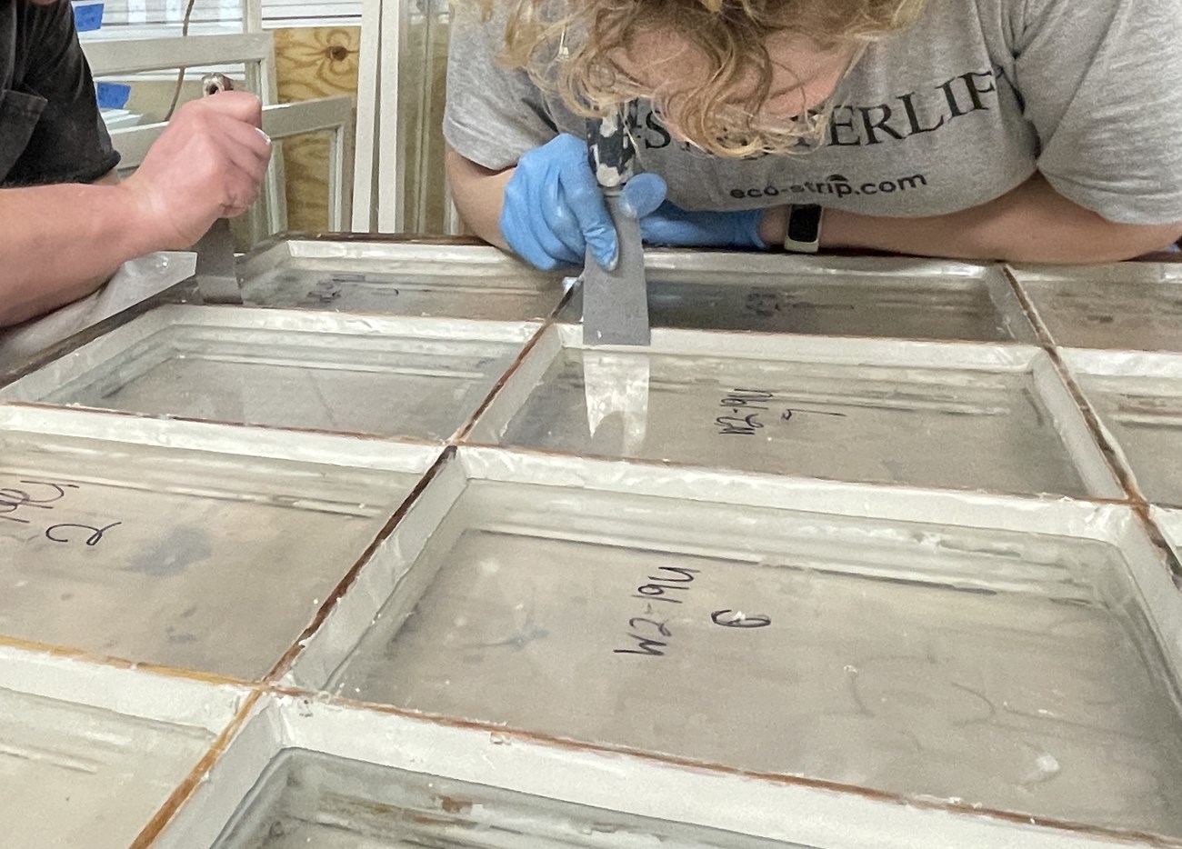 A closeup of a window with two crew members working on it. The window has freshly applied glaze on it, connecting the glass to the muntin bars. The crew members are each holding a putty knife and smoothing out the glaze.