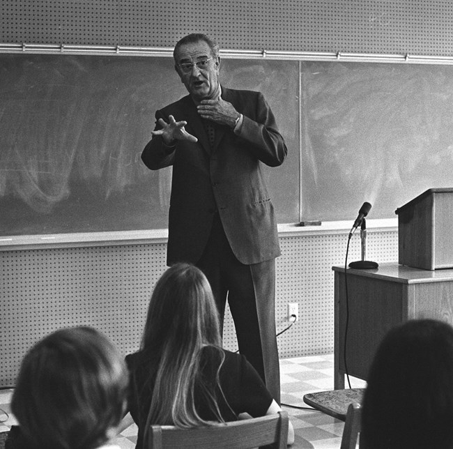 A man in a suit stands in front of chalkboards as he talks and gestures to a group of youth seated in desks.