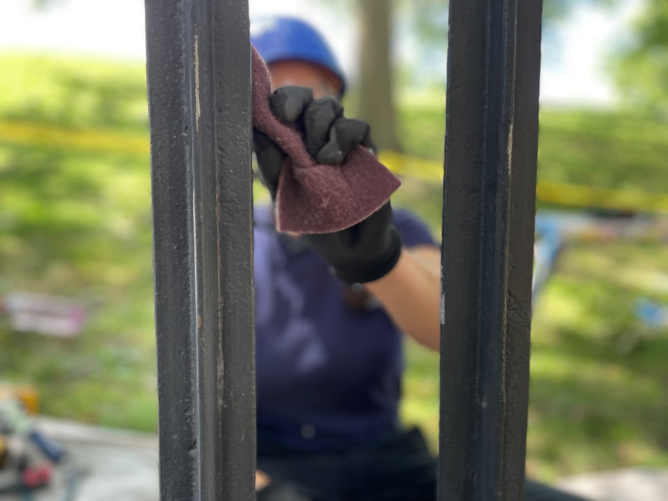 Closeup of two bars of an aged, wrought-iron fence painted black. In the background of the photo, out of focus, is a crew member wearing a hard hat. In the foreground is the crew member’s hand, wearing a glove and using a steel wire pad to brush the fen