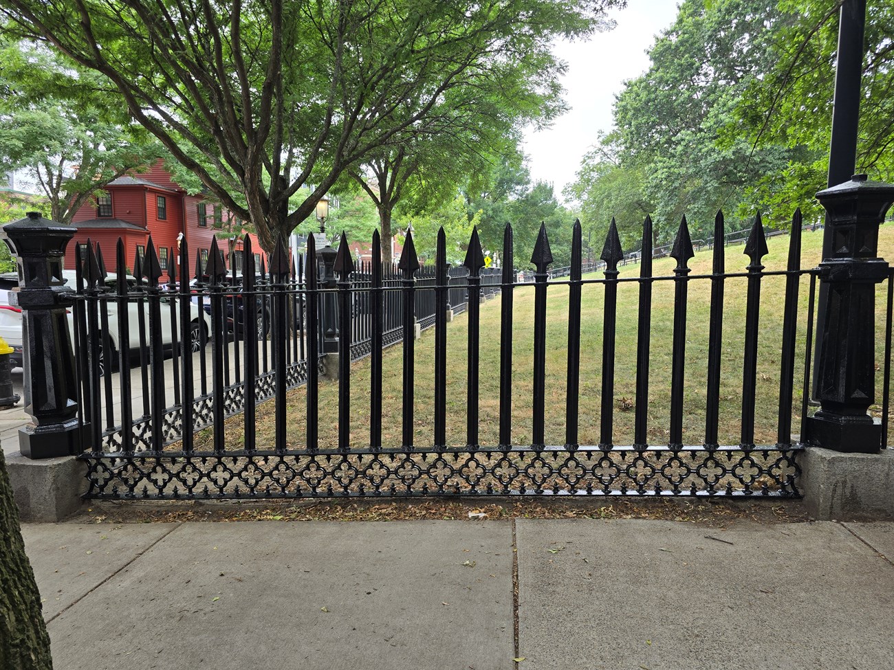 BHFimg5_fence Section of black wrought-iron fence. At each end is a decorative pillar, and there are 22 bars between the pillars. The tip of each bar alternates between a straight spike or triangular spike. The bottom section of the fence has a floral design.