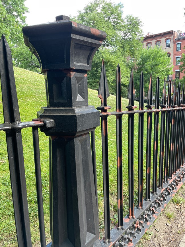 BHFimg6_primer2 A black wrought-iron fence from a side view. A thick pillar and several smaller bars of the fence are in view, with a floral pattern along the bottom of the fence. The fence has some spots of rust-colored paint applied to it.