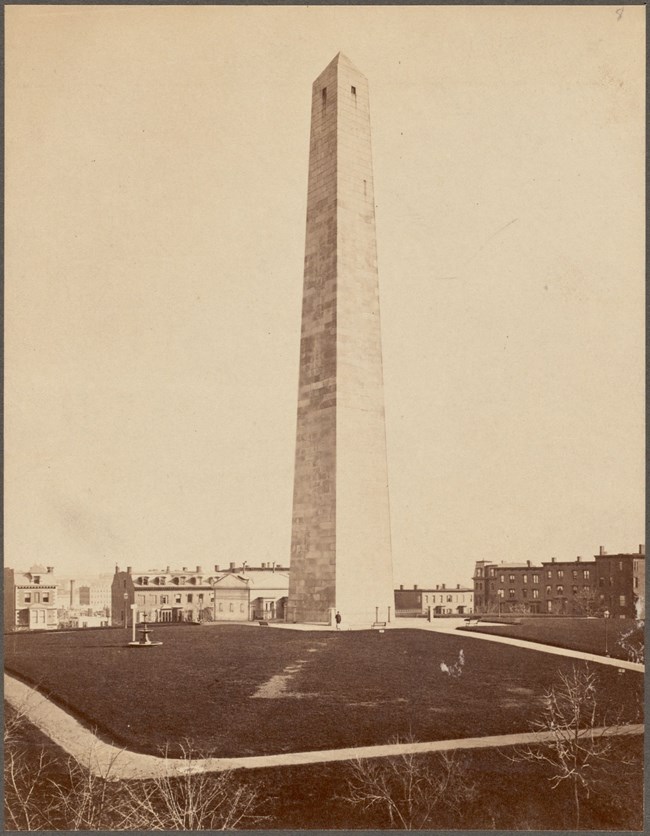 A black and white photo of the Bunker Hill Monument and Lodge on the top of a Hill.