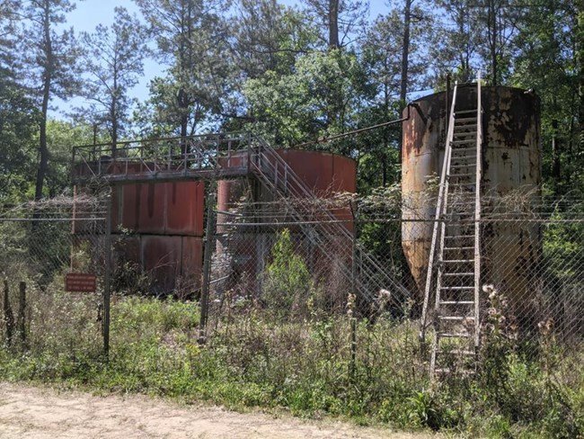 Three large tanks behind a fence with a bridge between the smaller tanks to the left and a ladder on the taller tank to the right