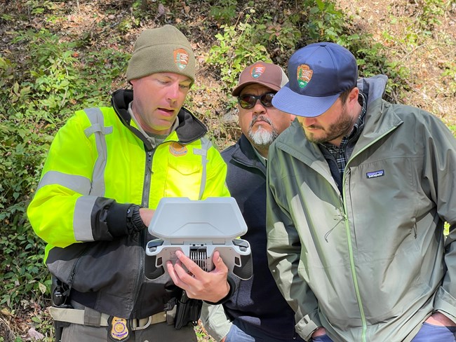Member of sUAS module shows Director Chuck Sams uncrewed aircraft dashboard for assessments. One man points at a console while two other men look at the console.