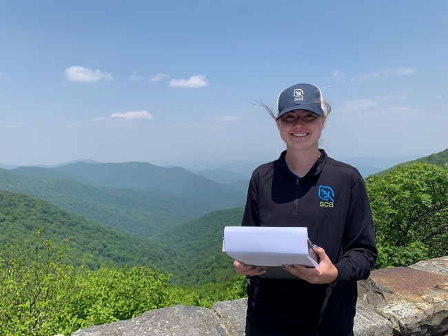 A woman wearing an SCA hat and polo stands outdoors with tree-covered mountains behind, holding a binder.