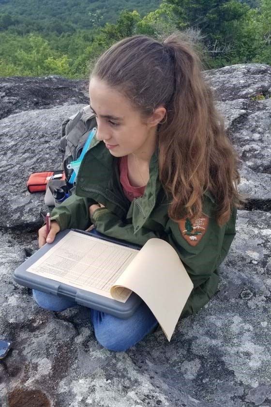 A woman in a green jacket sits on a rocky cliff while writing notes in her workbook
