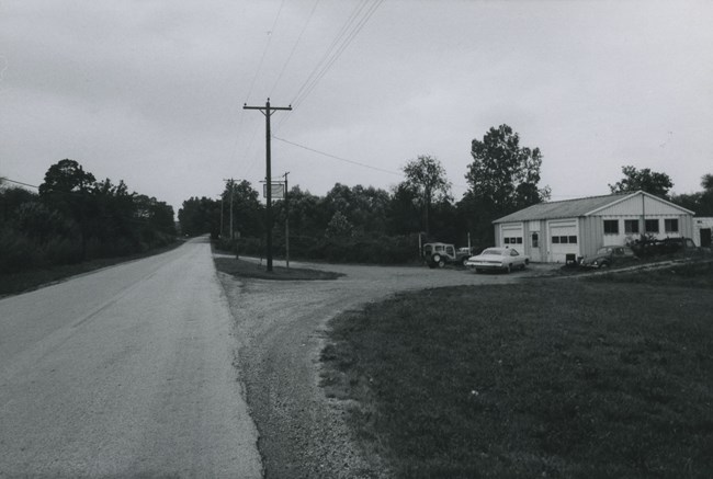 A two-bay garage stands along a rural road, marked by a sign hanging on a high pole.