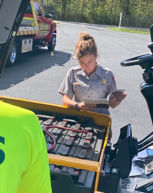 BMG New Forklift Female uniformed ranger stands looking down at a book, in front of the open hood of a yellow and black forklift.