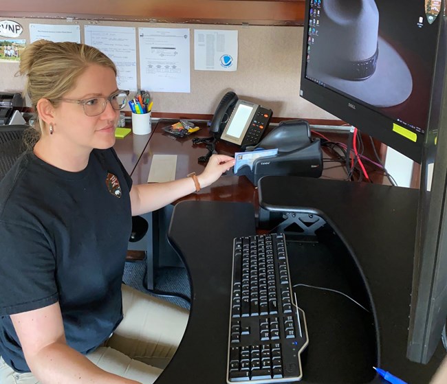 Female worker in blue shirt and khaki pants sits at a computer holding a check in a machine; the NPS arrowhead symbol appears on the screen and on her shirt.