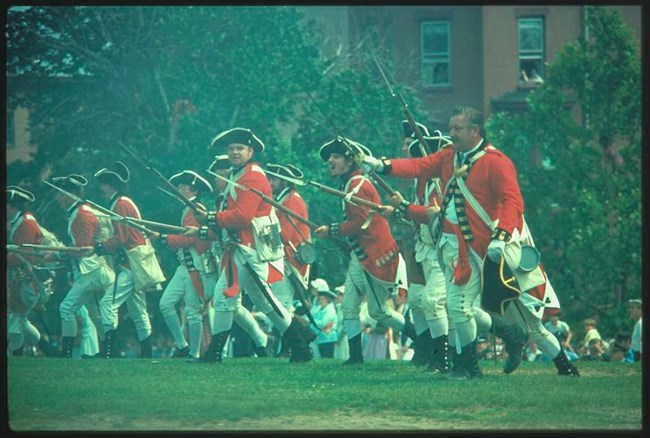 Reenactors in red coats charging up a grassy hill.