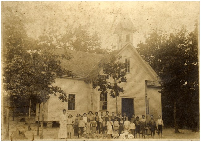 lt= White children standing outside a schoolhouse