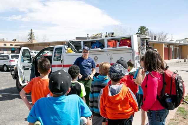 A group of about 9 elementary school students gather around a wildland fire engine, listening to a wildland firefighter talk.