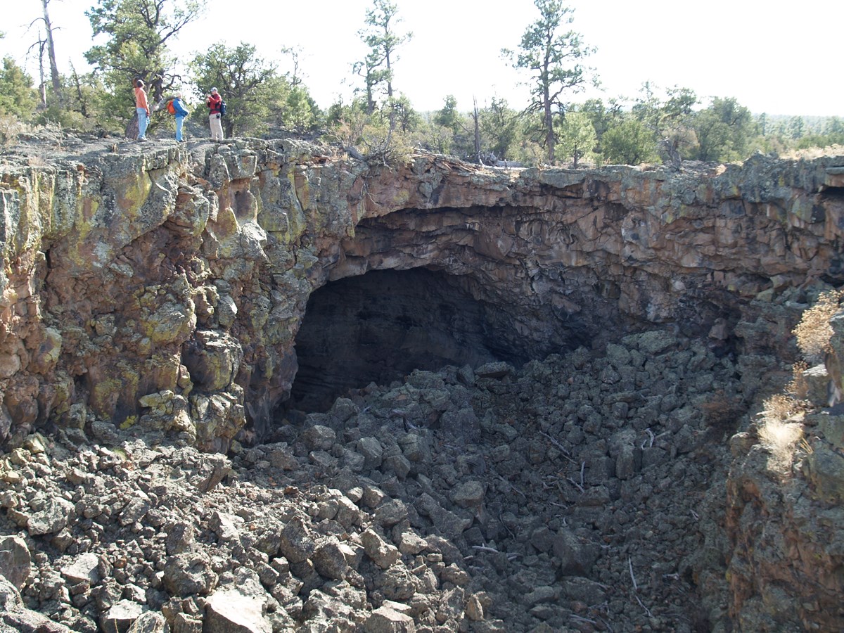 Photo of a rangers standing over a boulder covered pit with in a large lava tube cave opening in one wall