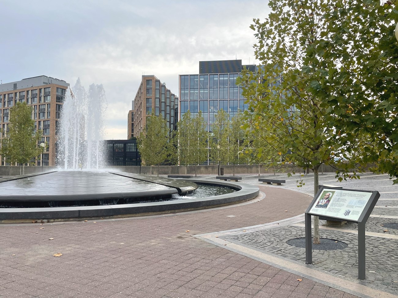Round fountain in the plaza of Benjamin Banneker Park with wayside information panel on right