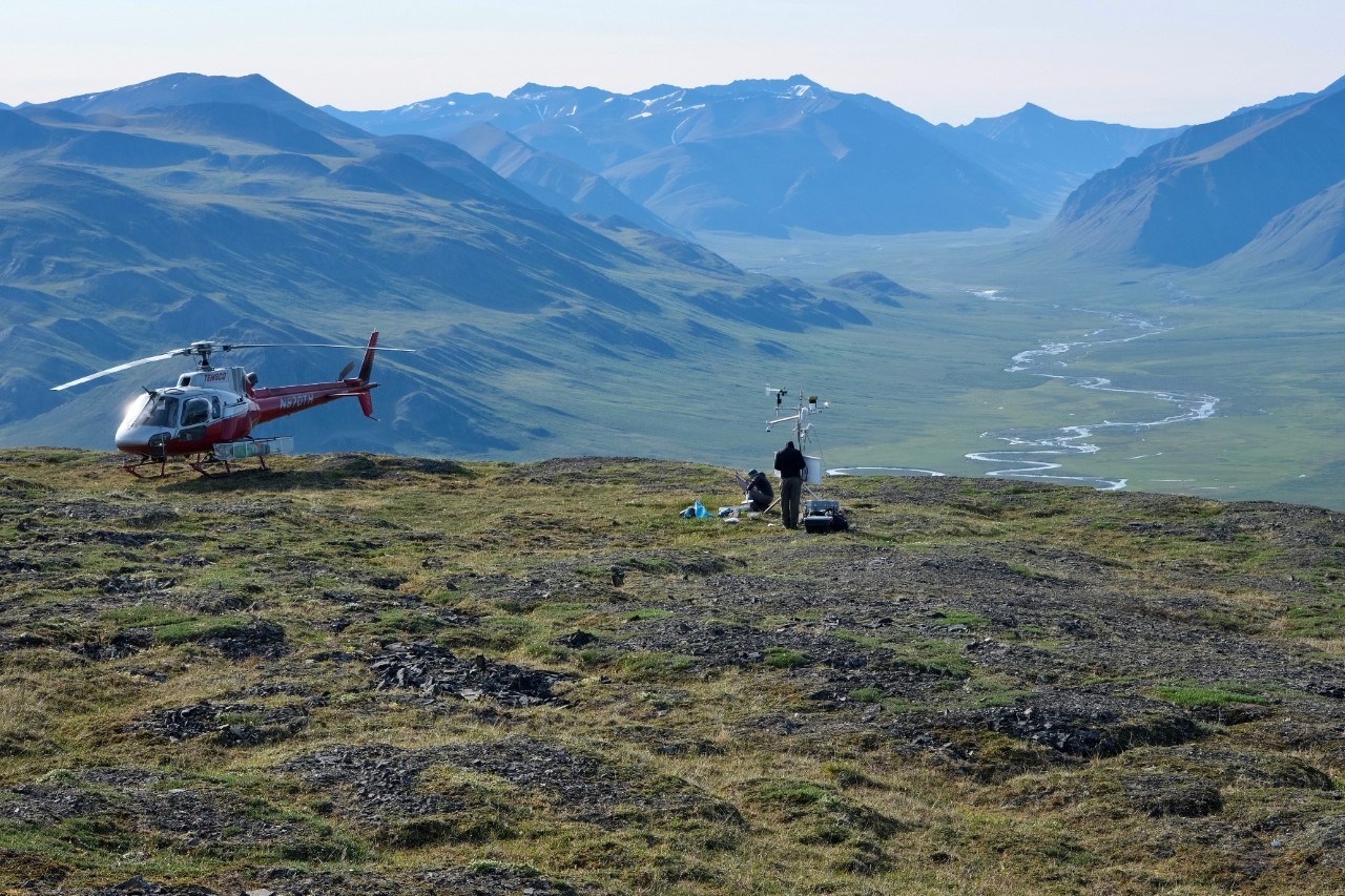Killik Climate Station in Gates of the Arctic National Park & Preserve A helicopter standby while two people work on a climate station on a bench perched above the Killik valley with mountains and a river in the distance