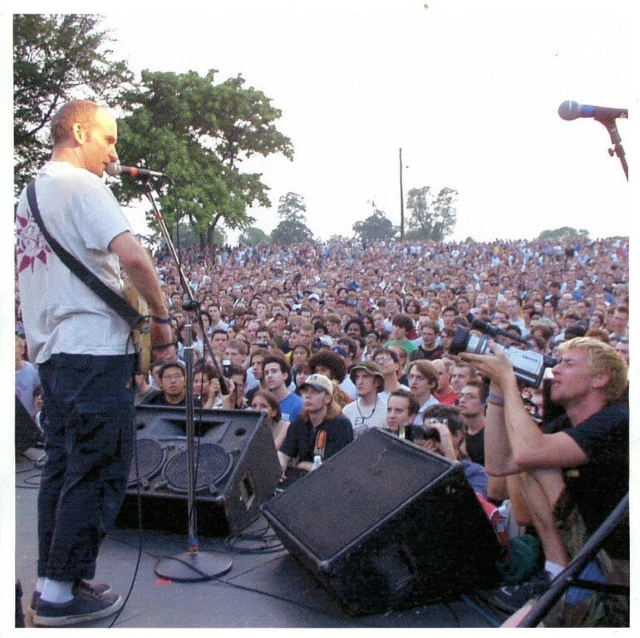 Fugazi performing at Fort Reno. July 1, 2002. As a man on stage plays guitar to a large audience, a camera man films him.