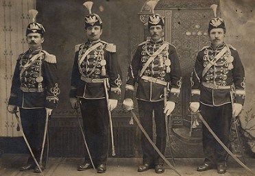 Photograph of four men in a line wearing feathered hats, regalia, and holding swords.