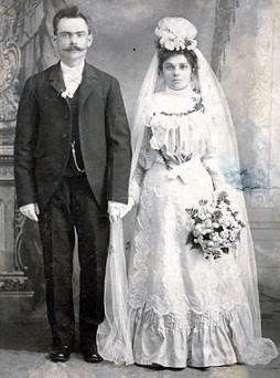Black and white wedding photo of a man in a suit standing next to his bride in a white lace gown.