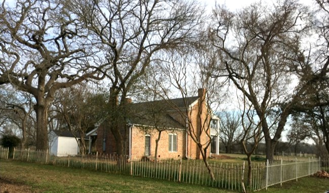 View of brick house surrounded by trees and a picket fence.