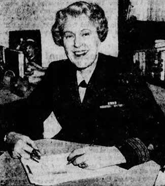 A woman wears a dark jacket with braiding at the cuffs.  She also has military ribbons pinned to her chest.  She holds a pen as she sits at a desk.