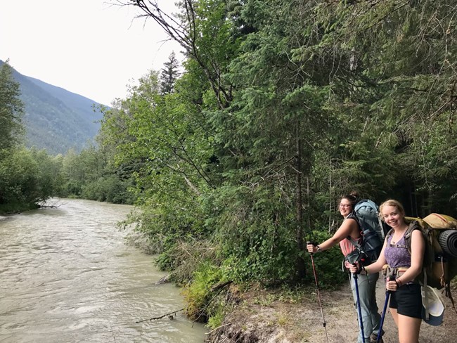 Two hikers stand on the bank of a river lined with trees.