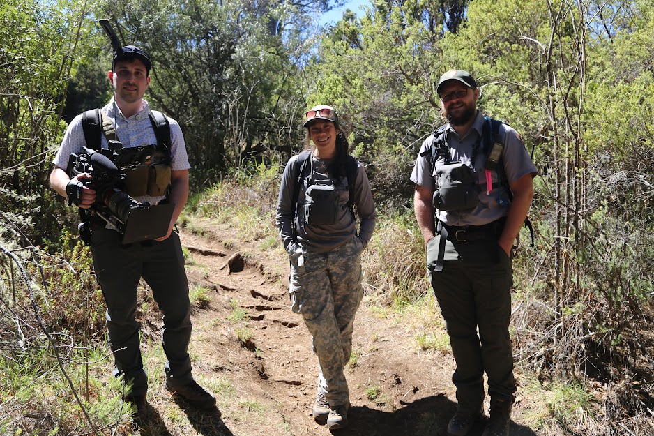 lt= Three people stand in a row on a dirt trail. One of them holds a large black camera with microphone attached.