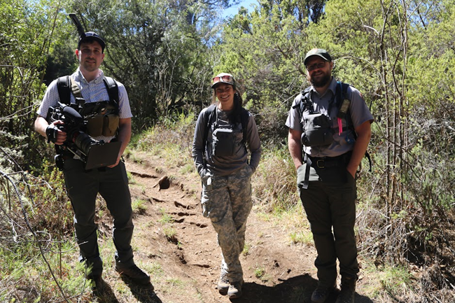 Three people stand in a row on a dirt trail. One of them holds a large black camera with microphone attached.