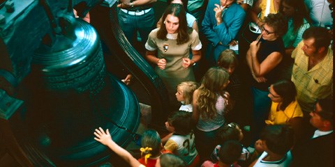 A visitor puts their hand on the Liberty Bell as a crowd surrounds a park ranger near the Liberty Bell.