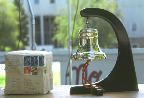 Color photo of small bell hanging from curved black stand, a small package is at left of bell