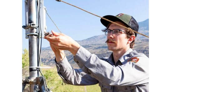 Man in NPS uniform working on equipment with grassy field, mountains, and blue sky in the background