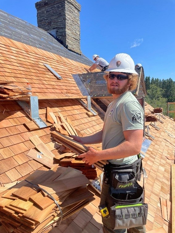 Ben Glowack Employee wearing sunglasses and white hardhat holds roofting tiles while working atop a building.