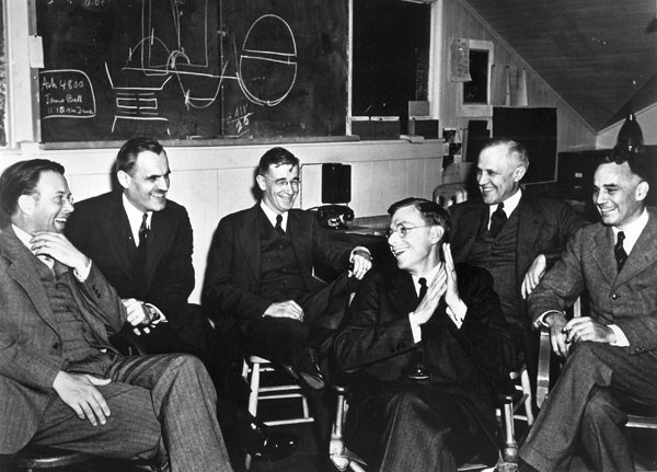 Black and white photo of six men in suits seated underneath a chalkboard.
