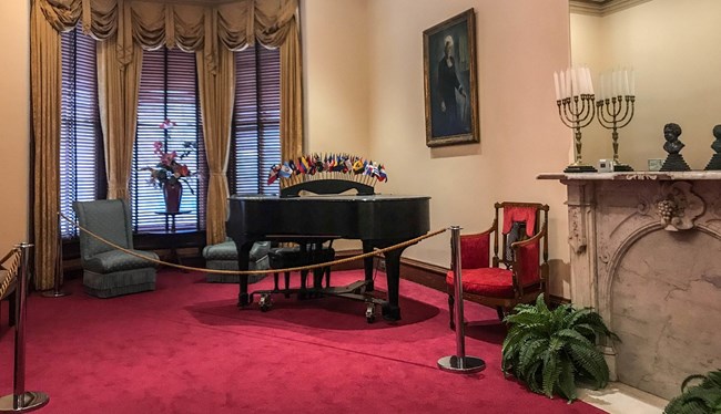 Parlor of a historic home that includes a portrail, piano covered in flags, and a fireplace