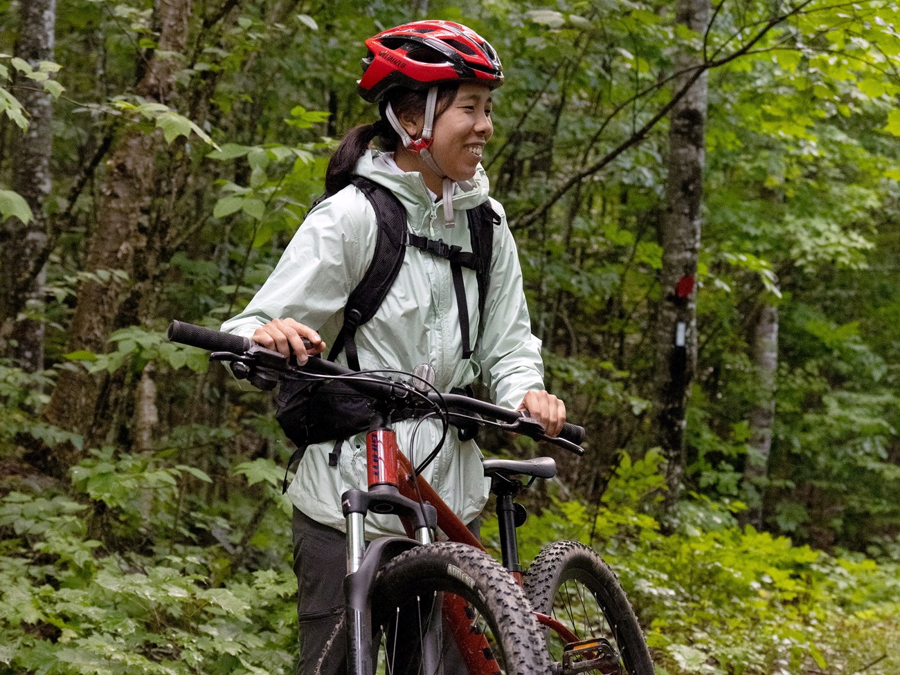 A woman stands smiling next to her red bike. She is wearing a red helmet and backpack. She is on an old logging road surrounded by green grass and foliage.