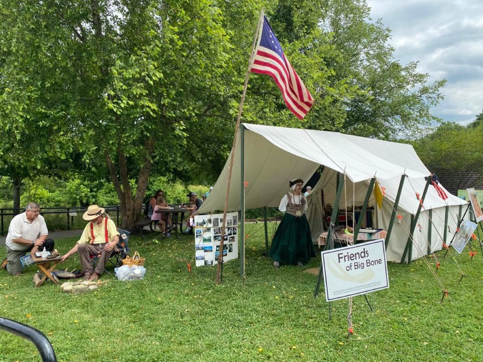 Around a canvas tent are two historical reenactors, and old U.S. flag, and a family on a picnic. Green grass and trees behind.
