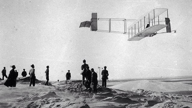 Black and white photo of a group of people standing on a beach watching a glider flying low in the sky.