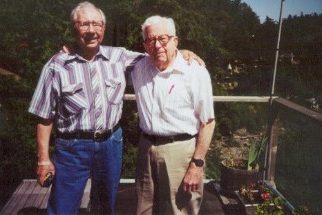 Color photo of two older men with white hair and glasses with their arms around each other.