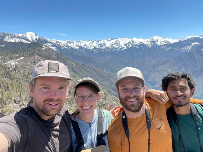 Selfie of three men and one woman wearing binoculars around their necks and smiling in front of spectacular mountain view.