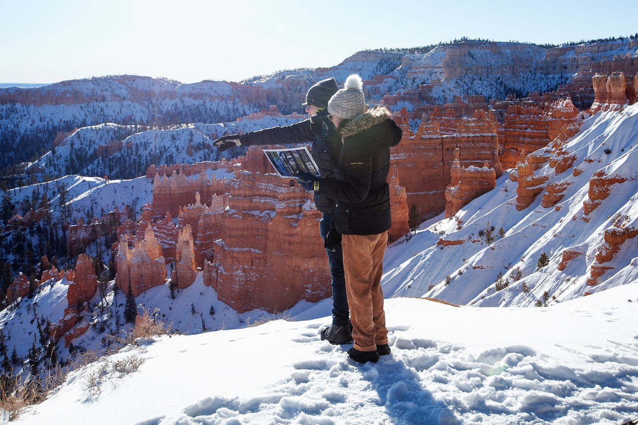 Birding at Edge of the Amphitheater in Bryce Canyon two people stand in snow above Bryce Canyon's Amphitheater pointing in the distance