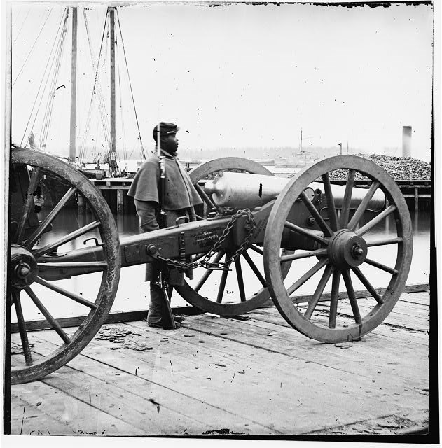 Black Civil War soldier standing guard, a cannon is positioned in front of him.
