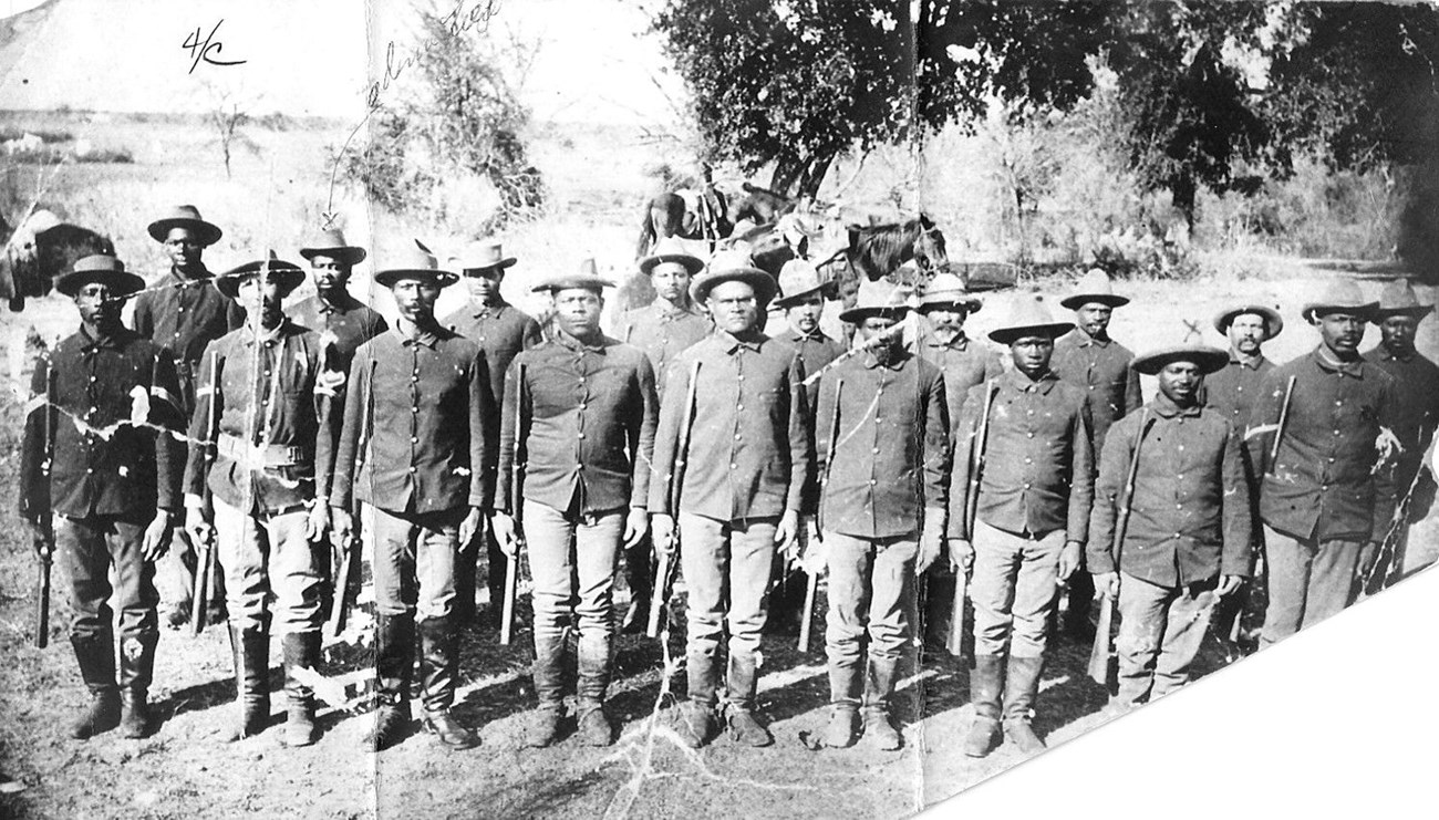 Black Seminole Indian Scouts circa 1885 Black and white phots of two rows of Black Seminoles standing at attention and looking directly at the viewer. They are all wearing the US Army uniform of the 1890s with black riding boots that come up to their knee. They are holding their carbines.
