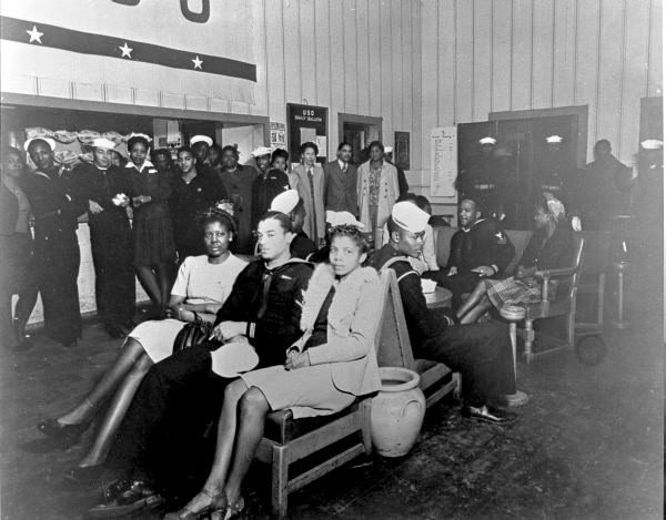 USO for African Americans, Pensacola Black and white photo of an African American man in uniform sitting between two African American women. African American men and women stand around the perimeter of the room, also in uniform
